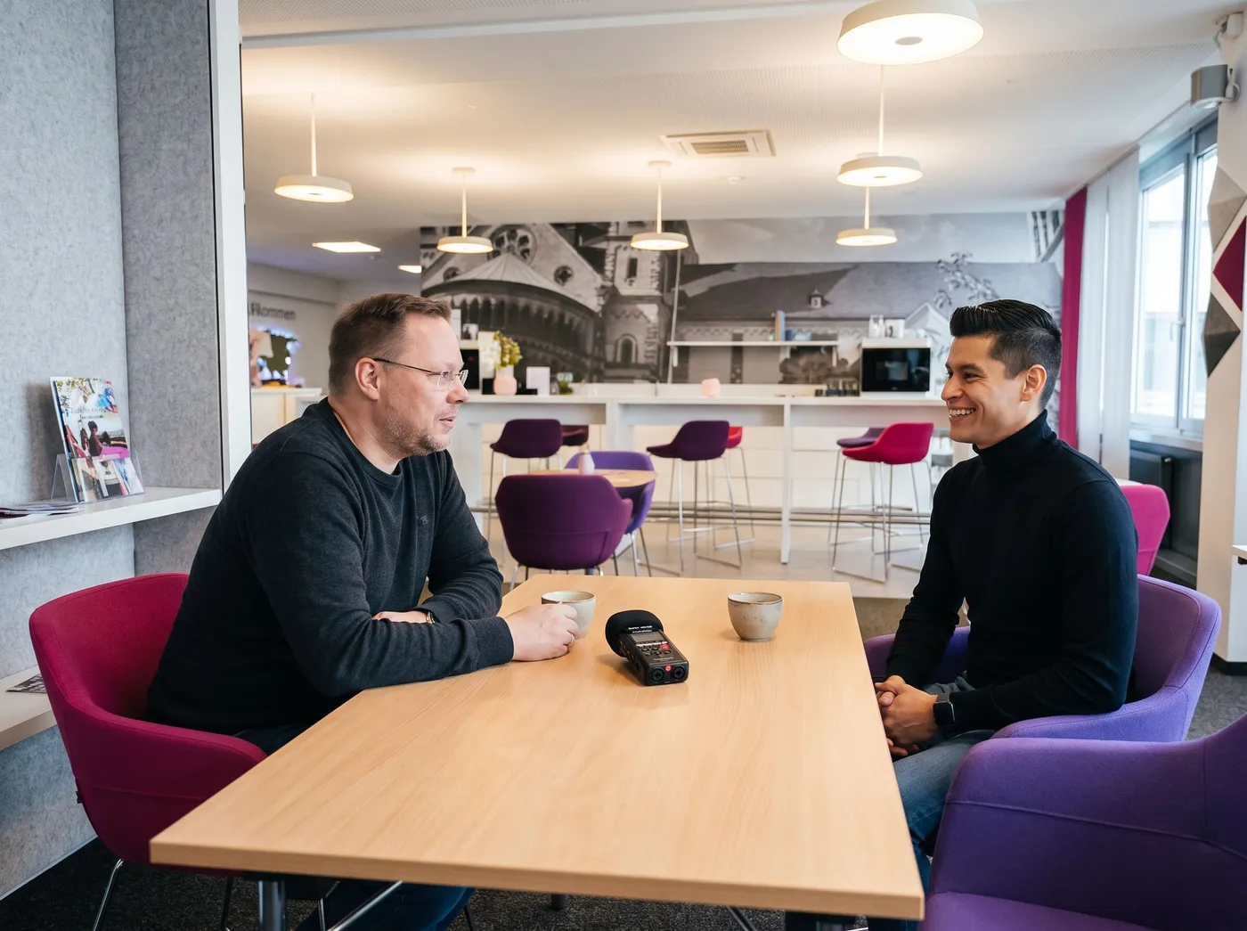 Stefan Haab und Marcel Avalos am Holztisch in Marcels Büro, in Magenta- und Lila-Sesseln, dazwischen das Aufnahmegerät